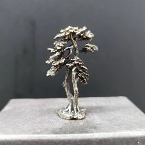 Puede incluir: Escultura metálica en miniatura de un árbol bonsái. El árbol tiene un aspecto detallado y texturizado con un acabado oscuro y envejecido. Las ramas y el follaje están intrincadamente elaborados, y el árbol está montado sobre una pequeña base.