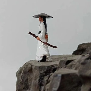 Op de afbeelding: Een klein beeldje van een persoon die een witte robe en een breedgerande hoed draagt, met een zwaard in de hand. Het beeldje staat op een rots.