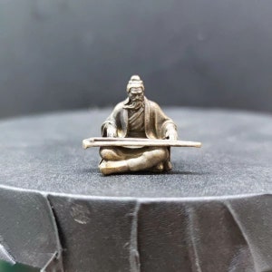 May include: A small silver figurine of a person playing a stringed instrument. The figurine is sitting in a meditative pose with their hands on the instrument. The instrument has a long neck and a rounded body.
