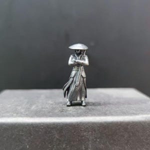 May include: A small, silver-colored figurine of a person in traditional attire, holding a staff and wearing a hat. The figure has crossed arms and stands on a dark surface. The figurine's details are finely crafted.