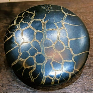 May include: A round, dark blue stone with a network of gold-colored veins. The stone has a smooth, polished surface.
