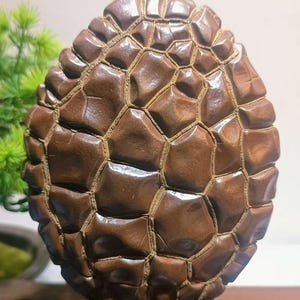 May include: A brown, egg-shaped sculpture with a textured, geometric pattern. The surface is divided into irregular, raised sections, creating a mosaic-like appearance. The sculpture is mounted on a small, dark brown wooden base.