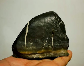 May include: A dark, irregularly shaped stone with white and yellow streaks. The stone is held in a hand, showcasing its natural texture and color variations. The stone's surface appears smooth and polished.