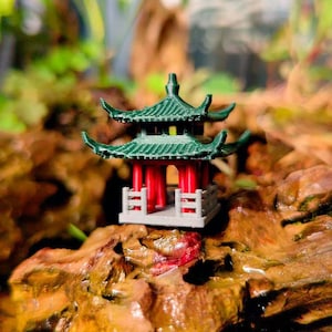 May include: A small, green, multi-tiered pagoda with red pillars and a gray base, sitting on a brown rock.