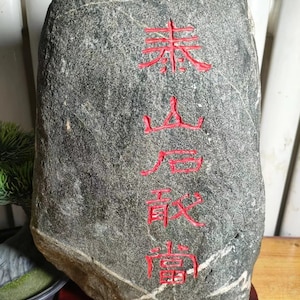 May include: A gray stone with red Chinese characters carved into it. The characters read "素心高致". The stone is sitting on a wooden stand.