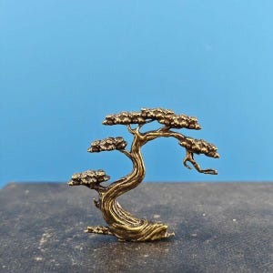 Könnte beinhalten: Eine kleine, goldfarbene Metallskulptur eines Bonsai-Baums. Der Baum hat einen dicken Stamm und viele Äste mit kleinen Blättern.