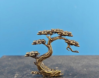 May include: A small, gold-colored metal sculpture of a bonsai tree. The tree has a thick trunk and many branches with small leaves.