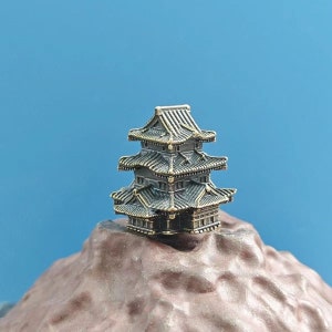 Puede incluir: Una pagoda miniatura de tres pisos hecha de latón con una pátina marrón oscura. La pagoda está sentada sobre una superficie rocosa marrón.