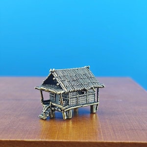 May include: A small, detailed model of a traditional house with a thatched roof, made of metal and painted in a dark brown color. The house is elevated on stilts and has a small porch with steps leading up to the entrance.