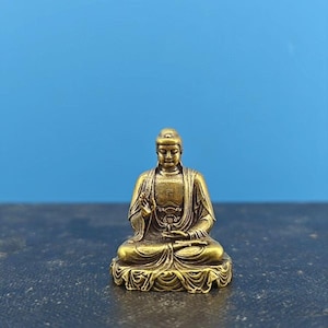 May include: A small, gold-colored Buddha statue in a seated meditation pose. The figure has a serene expression and is wearing a robe. The statue is on a decorative base, with a blue background.