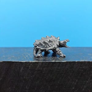 Könnte beinhalten: Detaillierte, dunkle Metallfigur einer drachenartigen Kreatur mit offenem Maul, stacheligem Rücken und vier Beinen. Die Figur ist vor einem blauen Hintergrund platziert.