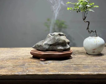 May include: A gray stone incense burner with smoke rising from it, sitting on a wooden base. The burner is on a wooden table with a small potted bonsai tree to the right.