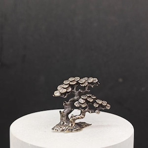 Puede incluir: Una pequeña escultura de metal de color plateado de un bonsái con una base texturizada. El árbol tiene muchas ramas y hojas.