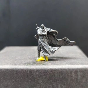 May include: A silver miniature figurine of a person wearing a long flowing cloak and holding a sword. The figurine is standing on a yellow base.