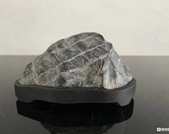 May include: A gray stone with a textured surface, displayed on a black base. The stone has a natural, organic shape and is a popular element in zen gardens.