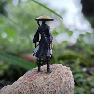 May include: A small figurine of a person wearing a black robe and a straw hat, standing on a rock. The figurine is holding a sword in their right hand.