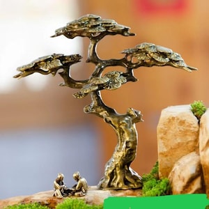 Puede incluir: Una escultura de árbol bonsái de bronce con hojas y ramas detalladas. Dos pequeñas figuras están sentadas en la base del árbol. La escultura está colocada sobre una pequeña formación rocosa con musgo verde.