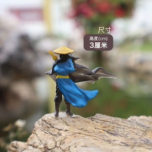 May include: A blue and black miniature figurine of a person wearing a yellow hat and holding a sword. The figurine is standing on a rock. The text "Size (cm) 3 centimetres" is visible in the image.