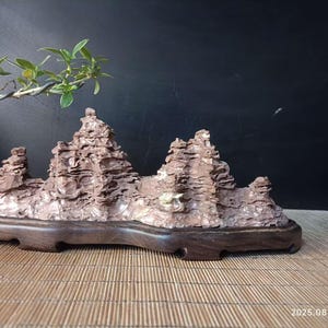 Op de afbeelding: Een miniatuur rotsformatie sculptuur met een donkerbruine houten basis. De sculptuur toont gelaagde, getextureerde rotsen in bruin- en beigetinten, die lijken op een bergketen. Een kleine bonsai-tak met groene bladeren is zichtbaar.