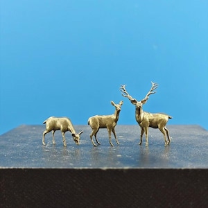 May include: Three golden deer figurines, one with antlers, standing on a black surface. The deer are facing different directions.