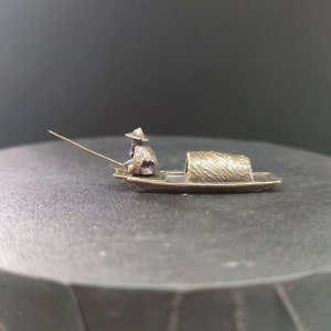 May include: A small silver figurine of a person in a boat with a fishing pole. The boat is made of metal and has a thatched roof. The figurine is detailed and realistic.