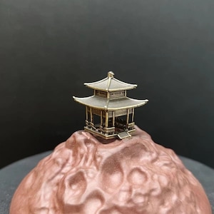 May include: A small, gold-coloured, two-tiered pagoda-style structure sits atop a textured, reddish-brown surface.