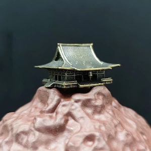 Puede incluir: Un pequeño modelo de bronce detallado de una casa japonesa tradicional con un techo a dos aguas que se asienta sobre una base texturizada de color marrón en forma de montaña.