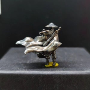 May include: A silver-coloured miniature figurine of a warrior wearing a wide-brimmed hat and a flowing cape. The warrior is holding a sword in their right hand and is standing on a yellow base.