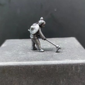 May include: A silver figurine of a person wearing a helmet and holding a tool. The figurine is standing on a grey surface.