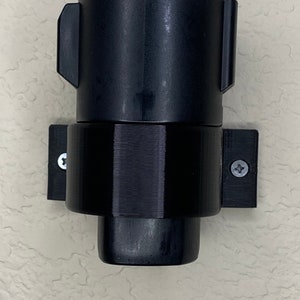 Puede incluir: Un soporte de pared de plástico negro para un cable de carga. El soporte tiene un tornillo a cada lado y un anillo texturizado alrededor del centro.