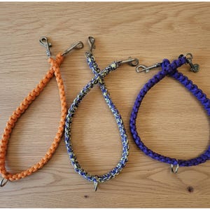 Op de afbeelding: Drie gevlochten hondenriemen in oranje, blauw en geel en paars. Elke riem heeft een metalen sluiting aan de bovenkant en een metalen ring aan de onderkant. De riemen worden gepresenteerd op een lichtbruin houten oppervlak.