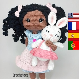 May include: A crocheted doll with brown skin and black curly hair is holding a white crocheted bunny wearing a pink dress. The doll is wearing a blue and pink dress.
