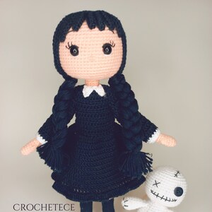 May include: A crocheted doll with black hair in pigtails wearing a black dress and white collar, holding a white crocheted doll with black stitching and a button eye. The dolls are on a white background. CROCHETETECE is written in the bottom left corner.