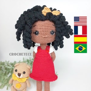 Könnte beinhalten: Gehäkelte Puppe mit brauner Haut, schwarzen lockigen Haaren und einem roten Kleid. Die Puppe hält einen kleinen gehäkelten Teddybär mit einer gelben Schleife.