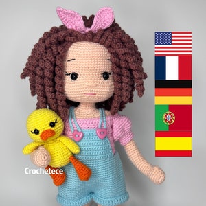 Könnte beinhalten: Eine gehäkelte Puppe mit braunen Locken, die ein rosa Shirt, eine blaue Latzhose und weiße Socken trägt. Die Puppe hält ein gelbes gehäkeltes Entchen. Die Puppe steht auf einem weißen Hintergrund. Der Text "Crochetece" ist im Bild sichtbar.