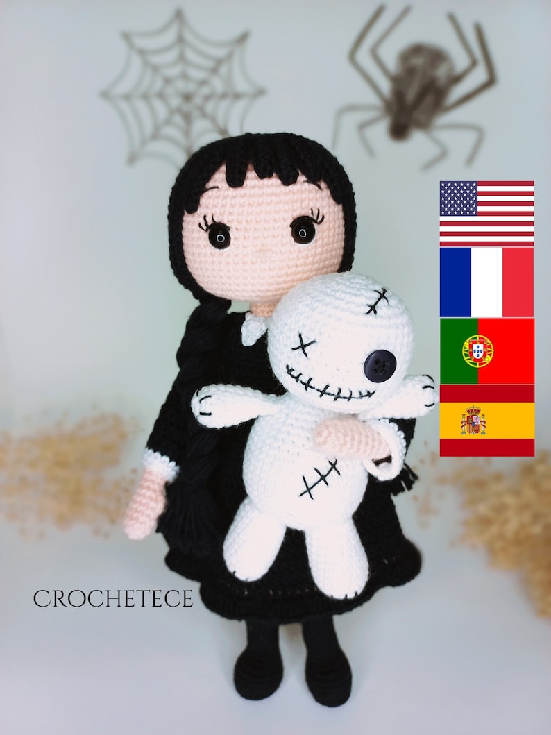 May include: A crocheted doll with black hair and a black dress is holding a white crocheted doll with stitched-on eyes and a stitched-on mouth. The doll is wearing a black dress and black shoes. The background is a white wall with a spider web and a spider.