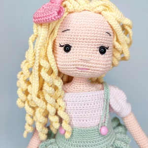 Könnte beinhalten: Eine gehäkelte Puppe mit blonden Locken, einer rosa Schleife und einem rosa-grünen Kleid. Die Puppe hat einen süßen Gesichtsausdruck und trägt ein rosa-grünes Kleid mit einem Rüschenrock.