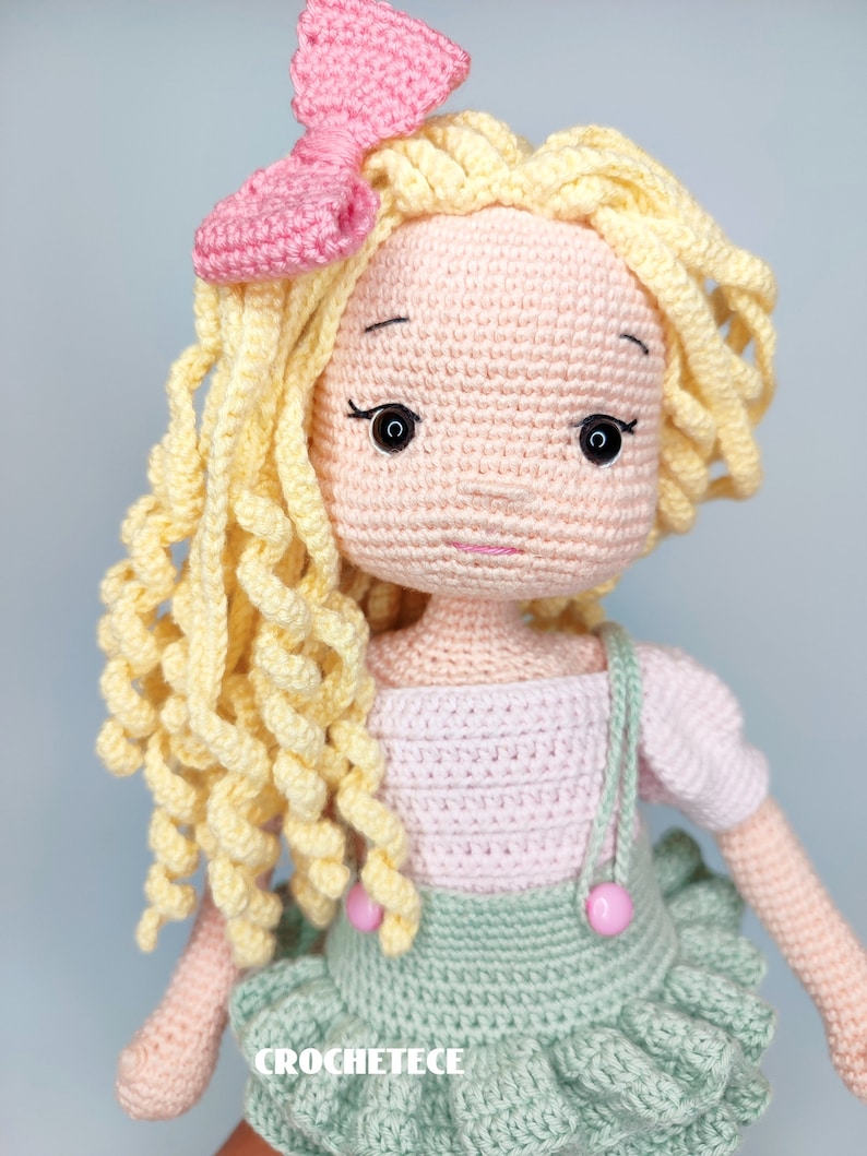 May include: A crocheted doll with blonde curly hair, a pink bow, and a pink and green dress. The doll has a sweet expression and is wearing a pink and green dress with a ruffled skirt.