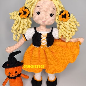 May include: A crocheted doll wearing an orange and black Halloween costume with a crocheted pumpkin friend. The doll has yellow hair with orange flower accents and black shoes.