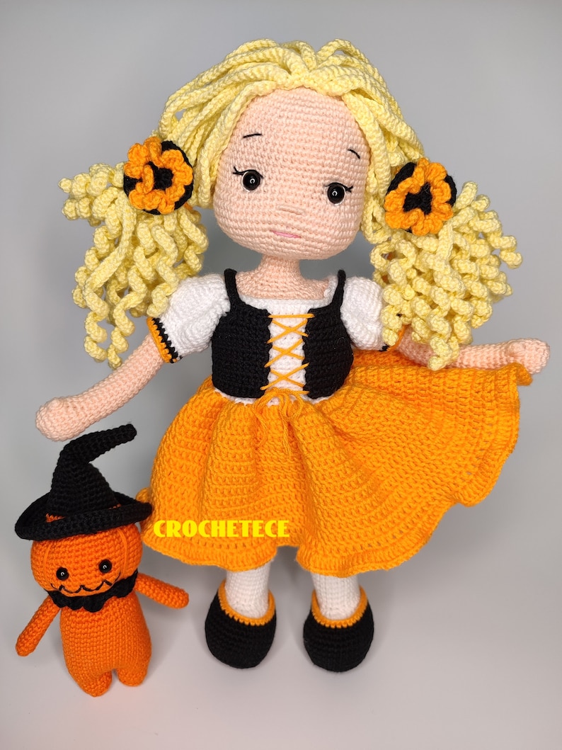May include: A crocheted doll wearing an orange and black Halloween costume with a crocheted pumpkin friend. The doll has yellow hair with orange flower accents and black shoes.