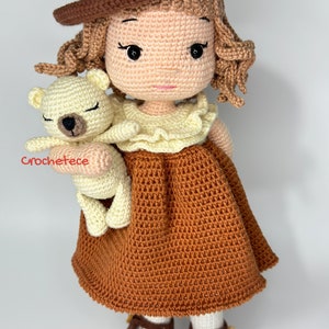 May include: A crocheted doll wearing a brown hat and dress, holding a crocheted white teddy bear. The doll has brown hair and brown eyes.