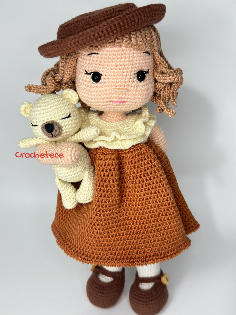 May include: A crocheted doll wearing a brown hat and dress, holding a crocheted white teddy bear. The doll has brown hair and brown eyes.