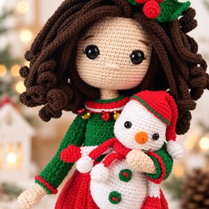 May include: A crocheted doll with dark brown curly hair, wearing a green sweater with small gold bells and a red skirt. The doll holds a small white snowman toy with a red hat, a red scarf, and two green buttons on its front. A green holly leaf with three red berries is attached to the doll's hair.