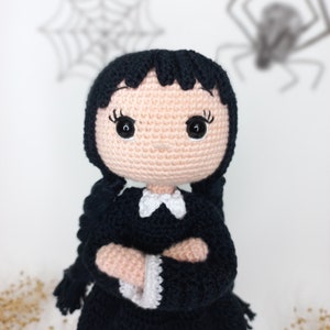 May include: Crocheted doll with black hair, a black dress, and a white collar. The doll has a pink face with black eyes and is standing in front of a white background with a spider web and a spider.