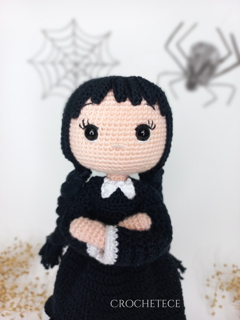 May include: Crocheted doll with black hair, a black dress, and a white collar. The doll has a pink face with black eyes and is standing in front of a white background with a spider web and a spider.