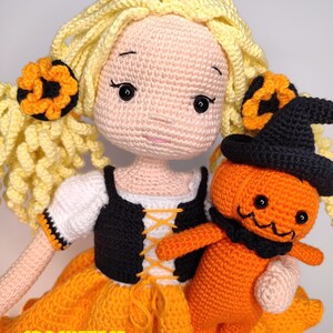 May include: A crocheted doll with blonde hair wearing an orange and black dress and holding a crocheted pumpkin doll. The pumpkin doll is wearing a black witch hat.