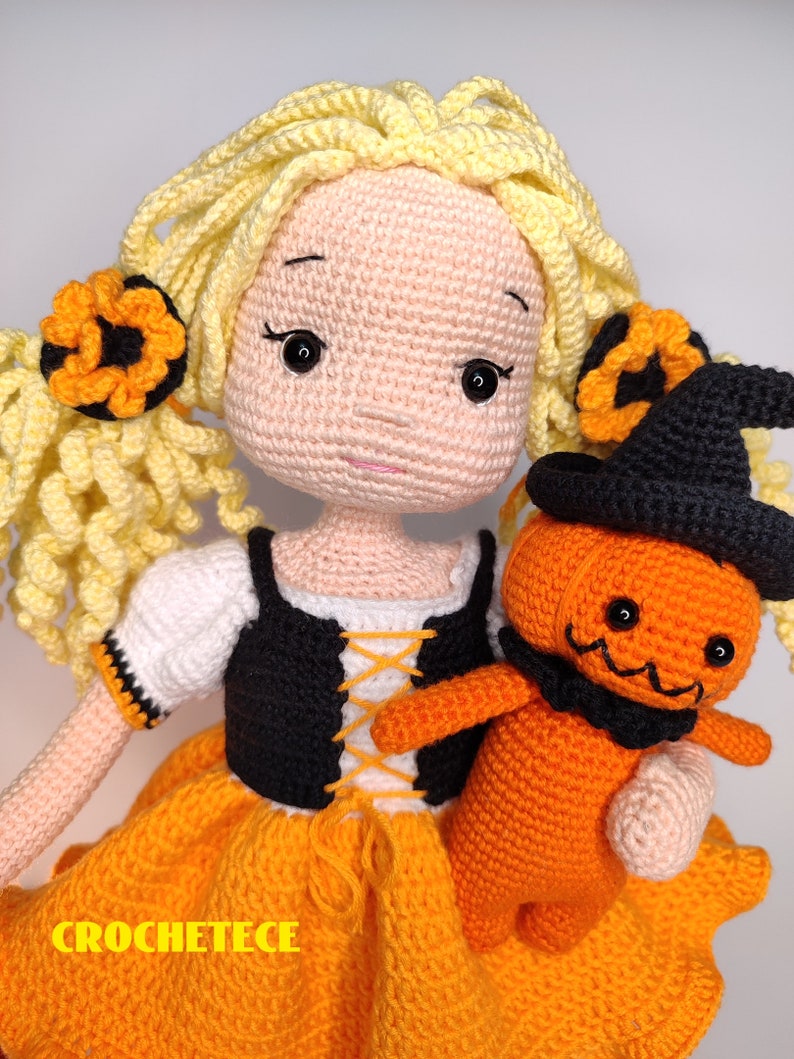 May include: A crocheted doll with blonde hair wearing an orange and black dress and holding a crocheted pumpkin doll. The pumpkin doll is wearing a black witch hat.