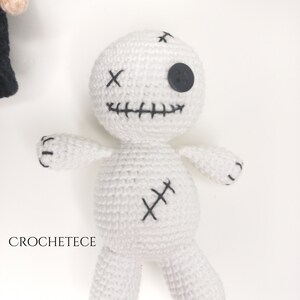 May include: White crocheted voodoo doll with black stitching and a black button eye. The doll has a stitched smile and a stitched X on its chest.