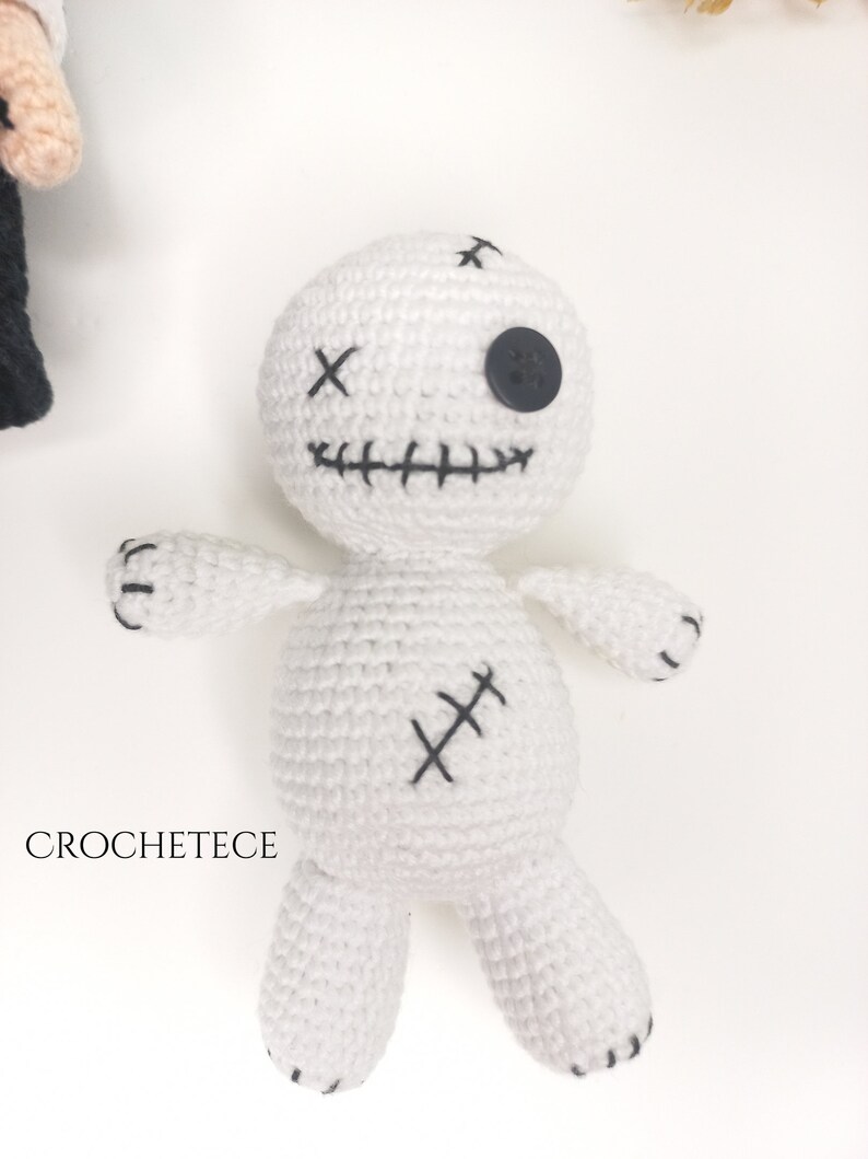 May include: White crocheted voodoo doll with black stitching and a black button eye. The doll has a stitched smile and a stitched X on its chest.