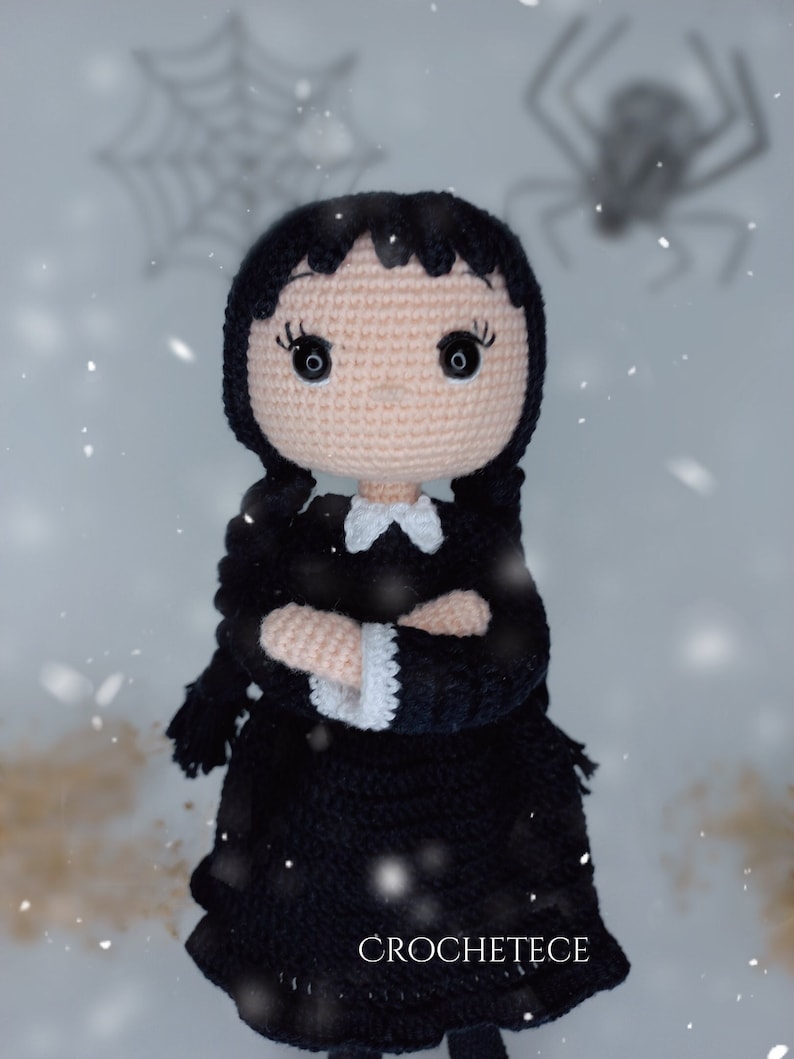May include: Crocheted doll with black hair, a black dress, and white collar. The doll has large black eyes and is standing in a snowy setting with a spider web in the background.